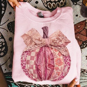 Peut inclure: Un sweat-shirt à col rond rose clair orné d'un grand motif de citrouille central. La citrouille est composée de divers empiècements en tissu dans des tons roses, incluant des motifs à carreaux, floraux et léopard, surmontée d'un nœud en dentelle.