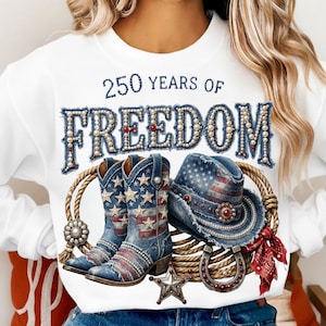 May include: White sweatshirt with a patriotic design featuring denim-style lettering that reads "250 YEARS OF FREEDOM." The design includes cowboy boots, a hat, a horseshoe, and a star, all with an American flag theme.