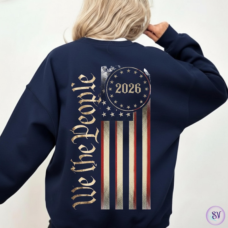 May include: Navy blue sweatshirt featuring a distressed American flag design. The flag includes red and white stripes, with a circle of stars and the year "2026". The words "We the People" are printed vertically on the left side in a gold color.