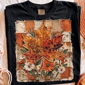 May include: A black t-shirt featuring a central square patch design with a large, detailed maple leaf in shades of orange, yellow, and brown. The background of the patch consists of a patchwork of textured fabric squares with floral accents.