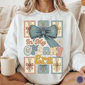 May include: White sweatshirt with a graphic design that reads "In My Granny Era." The design features a large blue bow, postage stamp-style graphics with bows, and text in various fonts and colors. A beige mug is visible in the image.