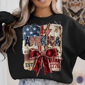 Peut inclure: Sweat-shirt noir avec un motif patchwork "AMERICA". Le motif comprend un champ d'étoiles bleu, des rayures rouges et blanches, des motifs floraux et un nœud rouge. Le texte "EST. 1776" est également visible sur le sweat-shirt.