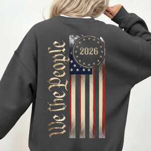 May include: Dark gray sweatshirt featuring a distressed American flag design. The flag includes red and white stripes, blue stars, and the year "2026" in a circle. The words "We the People" are printed vertically on the left side in a gold color.