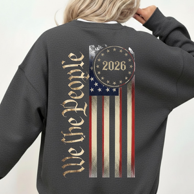 May include: Dark gray sweatshirt featuring a distressed American flag design. The flag includes red and white stripes, blue stars, and the year "2026" in a circle. The words "We the People" are printed vertically on the left side in a gold color.