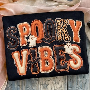 Könnte beinhalten: Ein schwarzes Sweatshirt mit dem Schriftzug SPOOKY VIBES in großen Stoffapplikationen. Die Buchstaben wechseln zwischen orangefarbenen, schwarzen und leopardenmuster-Stoffen. Kleine weiße Geister und schwarze Fledermäuse sind zwischen den Buchstaben aufgenäht.