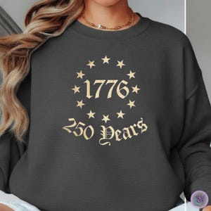 May include: Dark gray sweatshirt with a gold design featuring the year "1776" surrounded by stars, and the text "250 Years" in an old English font. The sweatshirt is a crew neck style.