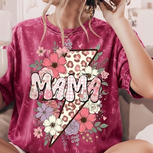 Peut inclure: T-shirt bordeaux avec le mot "MAMA" dans un design floral et à motifs. Le mot est placé sur un éclair avec un motif léopard. Le t-shirt est orné de fleurs roses, blanches et violettes.