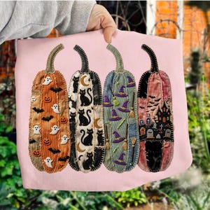 May include: A light pink sweatshirt featuring four tall, vertical pumpkin-shaped patches. The patches display embroidered Halloween motifs including ghosts, black cats, moons, witch hats, broomsticks, bats, and a haunted house with a graveyard.