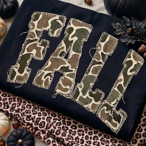 May include: A dark navy blue sweatshirt featuring the word FALL in large, block-style letters. Each letter is filled with a green, brown, and tan camouflage pattern and is outlined with visible, thick white decorative stitching.