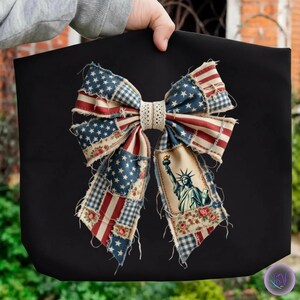 May include: Black tote bag featuring a large bow design with a patriotic theme. The bow incorporates elements of the American flag, including stars and stripes, along with an illustration of the Statue of Liberty.