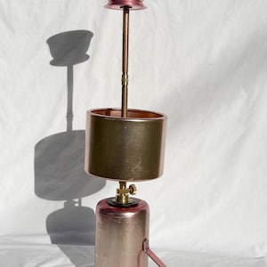 Puede incluir: Una lámpara de color cobre con una pantalla cilíndrica y un asa. La lámpara tiene una pequeña parte superior en forma de cuenco y un tallo largo y delgado. La base es más ancha y tiene un asa. La lámpara es de metal y tiene un acabado brillante.