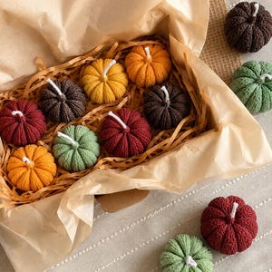Könnte beinhalten: Eine Sammlung kleiner, kürbisförmiger Kerzen in verschiedenen Farben, darunter Dunkelbraun, Salbeigrün, Dunkelrot und leuchtendes Orange. Mehrere Kerzen liegen in einer mit braunem Papier und Papierstreifen ausgelegten Pappschachtel.