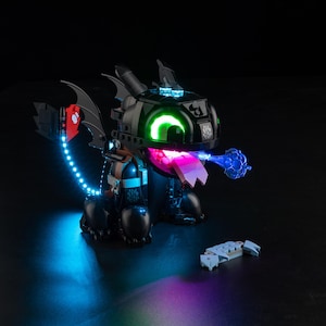May include: A black dragon toy with glowing green eyes and a pink tongue, emitting a blue light. The dragon has a red and white emblem on its wing and is surrounded by blue and purple lights. A small, detached piece sits nearby.