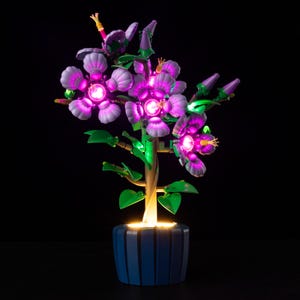 May include: A decorative illuminated flower arrangement in a blue pot. The artificial plant features purple and pink glowing flowers, green leaves, and a light-up base. The flower arrangement is set against a black background.