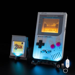 Op de afbeelding: Een lichtgrijze Nintendo Game Boy-replica met een verlicht scherm met een gepixeleerde scène. Het apparaat heeft een zwart kruisvormig richtingspad, roze knoppen en de tekst "Nintendo GAME BOY". Een spelcartridge met de tekst "ZELDA" is ook aanwezig.