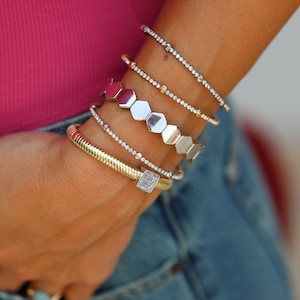 May include: A selection of bracelets in gold and silver. The image shows a gold, textured bangle, a silver bracelet with a square, diamond-like detail, and several delicate bracelets with small stones. Some bracelets have a hexagonal design.