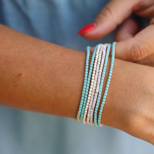 May include: Several delicate bracelets in shades of turquoise, white, and pale pink are stacked on a wrist. The bracelets are composed of small, evenly spaced beads, creating a layered look. The background is a soft blue.