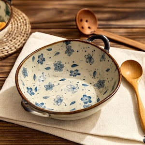 May include: A cream-coloured ceramic bowl with a brown rim, adorned with blue floral patterns and small handles. A wooden spoon is placed nearby. The bowl's design suggests a kitchen or dining setting, with a diameter of approximately 20 cm.