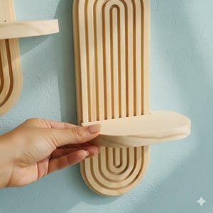 Op de afbeelding: Houten wandplank met een natuurlijke afwerking. De plank heeft een afgeronde bodem met gebogen lijnen, een platte plank en verticale lijnen erboven. De plank is gemaakt van lichtgekleurd hout en is gemonteerd op een lichtblauwe muur.