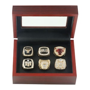 May include: A wooden display box with a glass top, showcasing six gold-toned championship rings. Each ring features intricate detailing, including gemstones and the word "Bulls". The box has a dark red finish and a black interior.