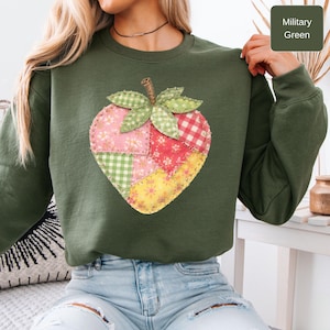 Puede incluir: Sudadera verde militar con un diseño de fresa de patchwork. La fresa está hecha de varias telas, incluyendo cuadros y estampados florales. La sudadera tiene mangas largas y cuello redondo. El texto "Military Green" está en la imagen.