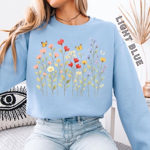 Könnte beinhalten: Hellblaues Sweatshirt mit einem Blumendesign aus roten, gelben und lila Blumen und Schmetterlingen. Der Text "LIGHT BLUE" ist vertikal auf dem rechten Ärmel aufgedruckt. Ein bequemer, lässiger Stil.