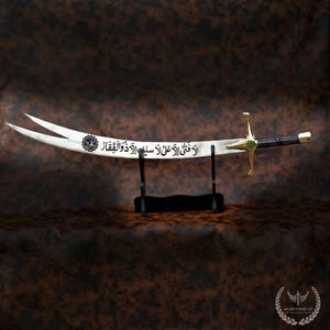 May include: A curved sword with a double-pointed blade, featuring gold accents on the hilt and a black handle. The blade has Arabic script and is displayed on a black stand. The sword is a decorative item.