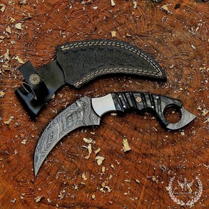 May include: A curved karambit knife with a Damascus steel blade and a black and wood-grain handle. The knife is paired with a black leather sheath with white stitching. The knife and sheath are displayed on a wooden surface.