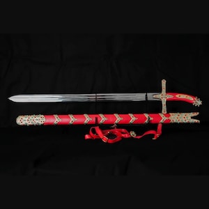 May include: A silver-toned metal sword with a curved red handle and a matching red scabbard. Both the hilt and the scabbard are decorated with gold-colored metal accents, small turquoise-colored stones, and red gems. A red strap is attached to the scabbard.