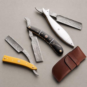 May include: Three straight razors with patterned Damascus steel blades and distinct handles in yellow, black, and white. The black handle features gold-tone pins and a central decorative inlay. A brown leather sheath is positioned to the side.