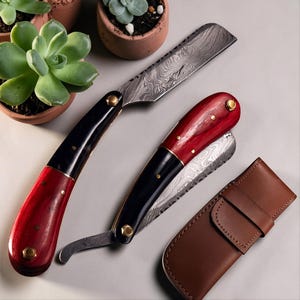 May include: Two straight razors with Damascus steel blades and handles featuring alternating sections of polished black and deep red material, secured with brass pins. A brown leather sheath with a flap closure sits beside the razors.