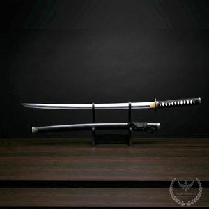 May include: A katana sword with a silver blade and black handle, displayed on a black stand. The scabbard is black and rests below the sword. The handle has a diamond pattern. The Valor Forge Co. logo is in the bottom right corner.