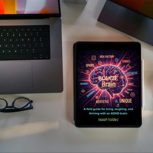 May include: A tablet displaying the book cover of "Bougie Brain" with a neon brain graphic. The cover text includes "A field guide for living, laughing, and thriving with an ADHD brain." A laptop, glasses, and mouse are also visible.