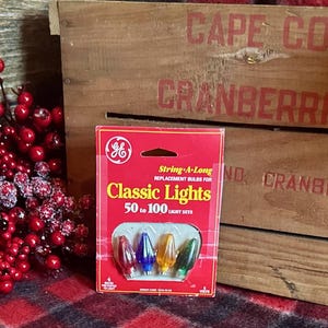 Puede incluir: Una caja roja de bombillas de repuesto GE Classic Lights. La caja presenta el texto "Classic Lights" y muestra cuatro bombillas de colores: rojo, azul, amarillo y verde. El fondo incluye una caja de madera con "Cape Cod Cranberries" impreso.