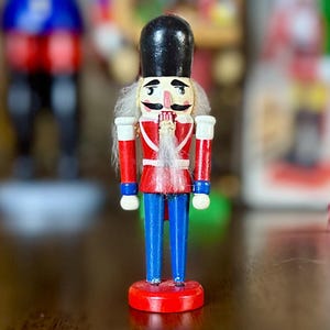 May include: A wooden nutcracker figurine, painted in red, blue, and black, stands on a red circular base. The figurine has a white beard and moustache, and is wearing a black hat. The background is blurred, suggesting other similar figurines.