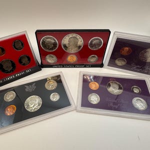 May include: Multiple United States Proof Sets displayed in protective cases. Each set contains various coins, including silver dollars, half dollars, quarters, dimes, nickels, and pennies. The sets are in black, red, and purple cases, with the words "United States Proof Set" visible.