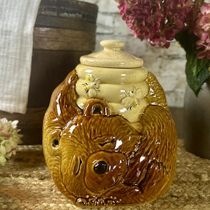 May include: A glossy ceramic honey jar featuring a sculpted brown bear hugging a cream-colored beehive. The beehive lid is topped with a small knob and decorated with two small painted bees. The bear has a smooth, shiny finish and dark eyes.