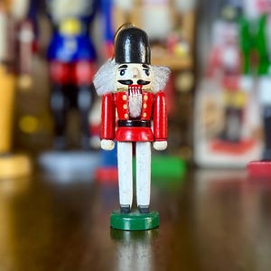 May include: A wooden nutcracker figurine with a red jacket, white trousers, and a black hat. The figurine has a white beard and moustache, and is standing on a green base. Other nutcrackers are visible in the background.