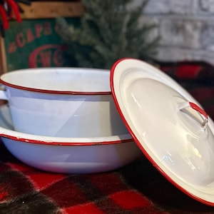 Może przedstawiać: Biała emaliowana miska i pokrywka z czerwonym obramowaniem. Miska ma dwa uchwyty i stoi na pasującym talerzu. Pokrywka jest lekko uchylona, ​​odsłaniając wnętrze. Przedmioty znajdują się na czerwono-czarnej kraciastej narzucie.