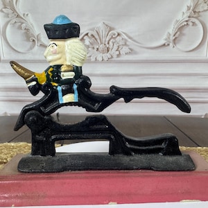 May include: A cast iron nutcracker shaped like a soldier. The soldier has a blue hat, blonde hair, and a painted face. The nutcracker is black with gold accents and sits on a red wooden base. The design is vintage and the measurements are in cm.