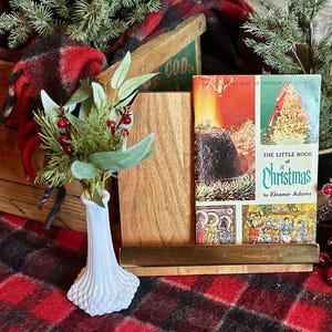 May include: A vintage Christmas scene featuring a book titled "The Little Book of Christmas" by Eleanor Adams, displayed in a wooden book stand. A white vase with greenery and red berries sits in front of the book. The background includes a plaid blanket and a wooden crate.
