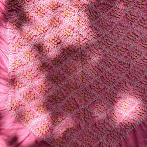 May include: A close-up shot of a pink fabric with a detailed pattern. The pattern features a grid of white lines with small, yellow-gold accents. The fabric has a textured, gathered appearance, with visible folds and shadows adding depth.