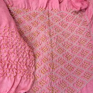 May include: Close-up of a pink textile with intricate embroidery. The fabric features a diamond pattern created with white thread and accented with small yellow stitches. The texture is soft and slightly ruffled, suggesting a delicate material.