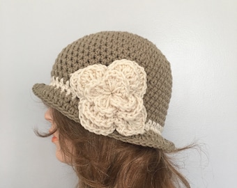 May include: A crocheted cloche hat in a muted olive green color, featuring a cream-colored flower applique. The hat has a small brim and a cream-colored band around the base. This handmade hat is a stylish accessory.
