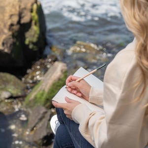 May include: A person writing in a notebook with a pencil. The notebook is open, revealing lined pages. The person is wearing a cream-colored hoodie and blue jeans. The background includes rocks and water.