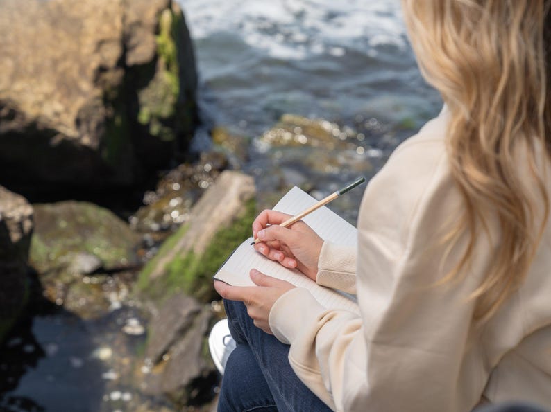 May include: A person writing in a notebook with a pencil. The notebook is open, revealing lined pages. The person is wearing a cream-colored hoodie and blue jeans. The background includes rocks and water.