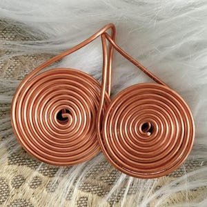 May include: Copper wire earrings featuring a spiral design. The earrings have a teardrop shape with a circular coil at the bottom. The copper has a warm, reddish-brown hue. The earrings are displayed on a white, fluffy surface.