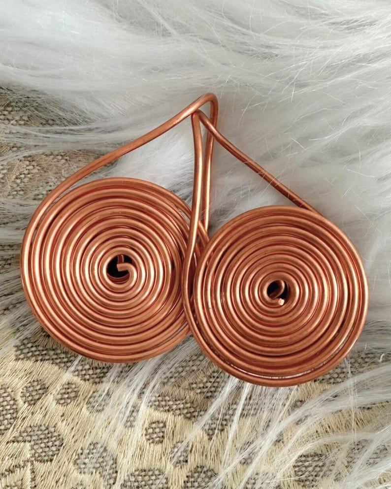 May include: Copper wire earrings featuring a spiral design. The earrings have a teardrop shape with a circular coil at the bottom. The copper has a warm, reddish-brown hue. The earrings are displayed on a white, fluffy surface.