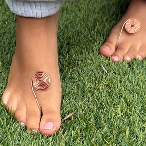 May include: Close-up of two feet with copper wire toe rings and ankle spirals. The spiral jewelry is on a background of green grass. The person is wearing gray sweatpants.