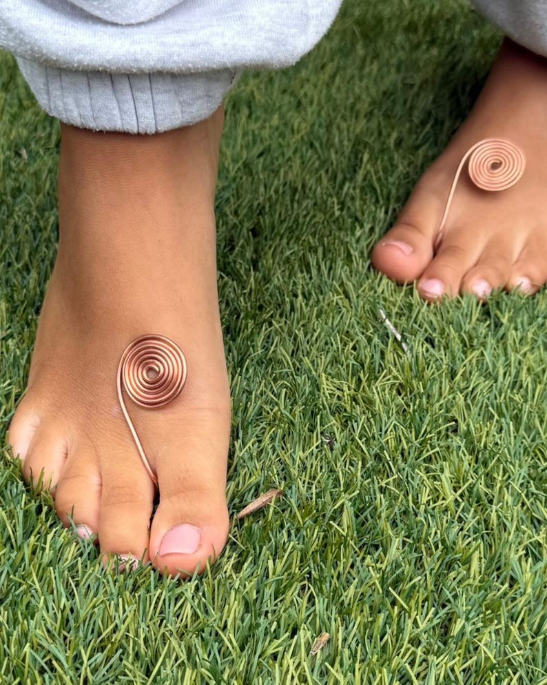 May include: Close-up of two feet with copper wire toe rings and ankle spirals. The spiral jewelry is on a background of green grass. The person is wearing gray sweatpants.
